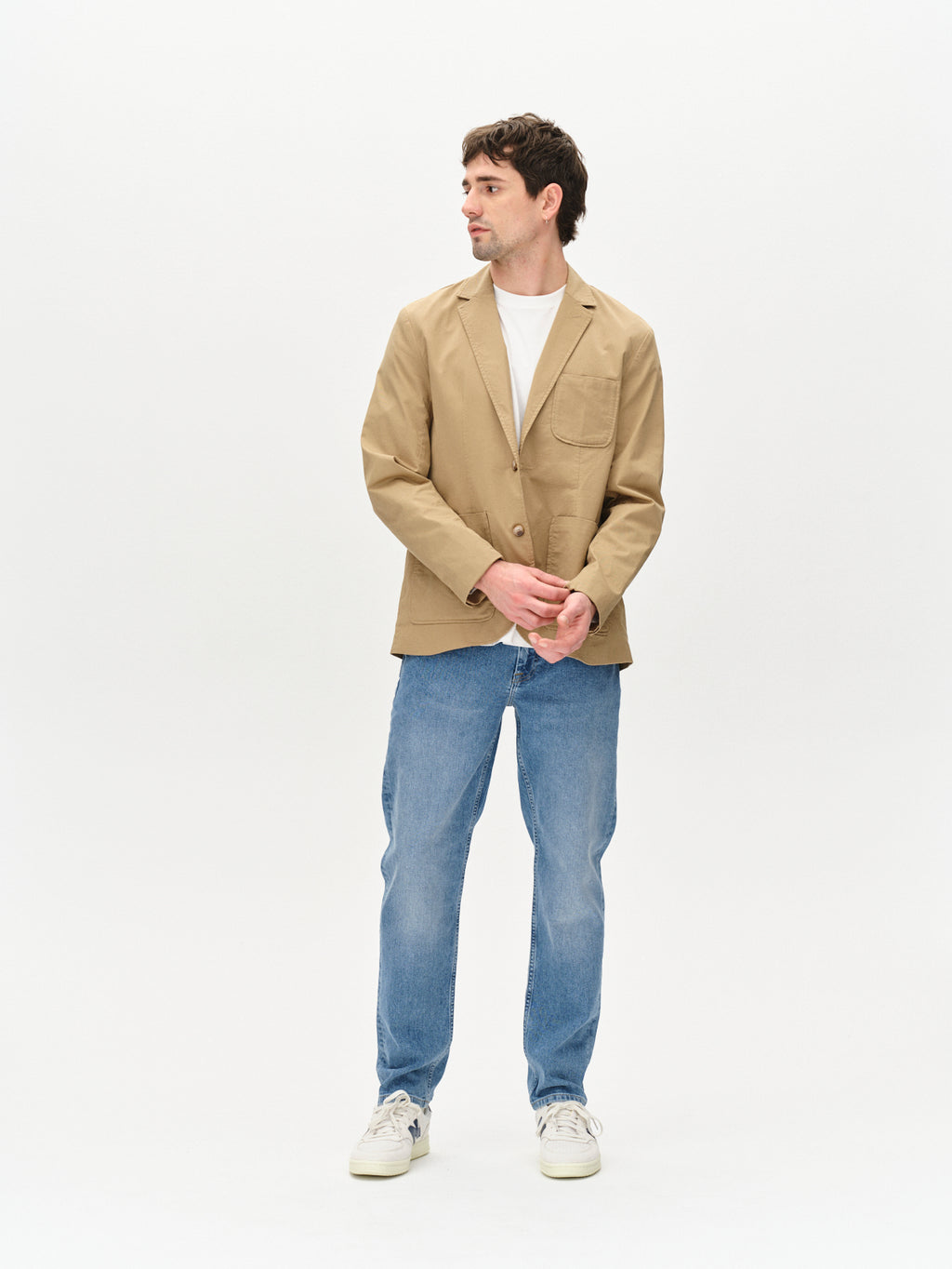 A man stands against a white background, wearing the Gabba Josh Unite - Sand blazer over a white t-shirt, blue jeans, and white sneakers. He looks to his left with his hands loosely clasped in front of him.