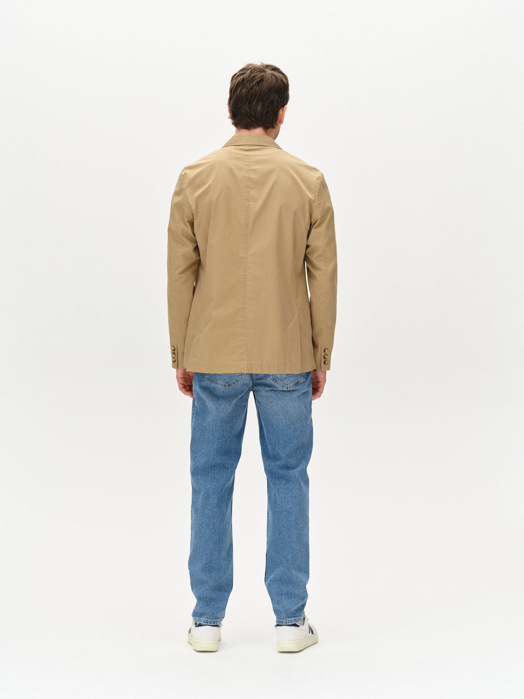 A person faces away from the camera wearing Gabbas Josh Unite - Sand blazer, light blue jeans, and white sneakers against a plain white background.