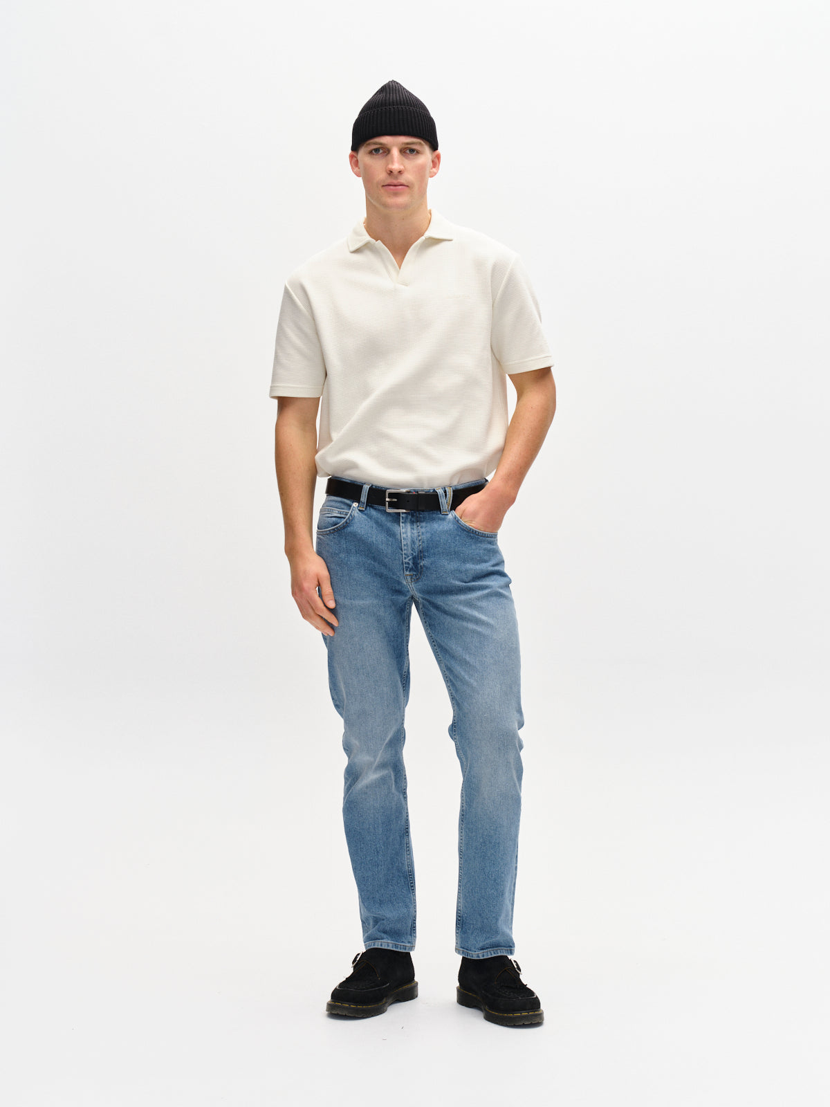 A man stands against a plain white background, wearing a black beanie, short-sleeve white polo, Gabbas Marc 5poc - Mid Blue Denim, and black shoes. One hand is in his pocket while the other rests by his side, capturing a modern classic look.