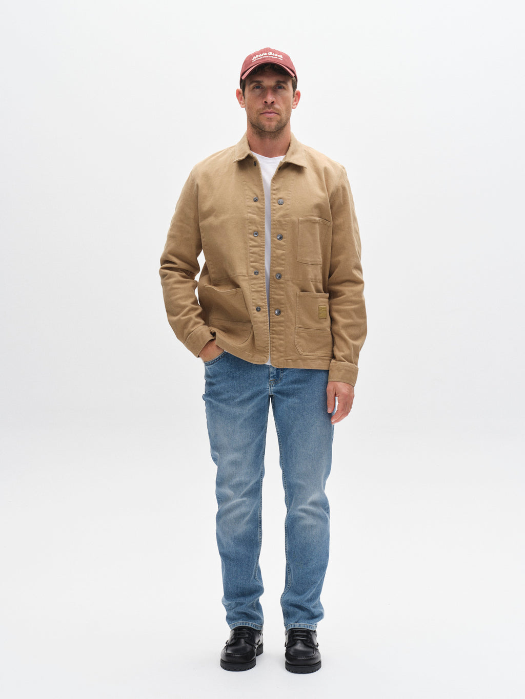 A man faces forward wearing a red cap, tan button-up jacket, white shirt, Gabba GABMarc 5poc - Mid Blue Denim jeans, and black shoes against a plain white background.