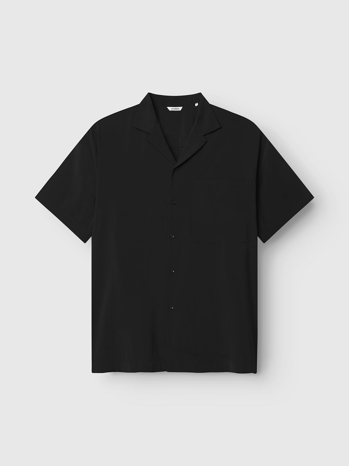 The Gabba Jens Over Resort Yari SS - Meteorite is a black short-sleeve button-up premium shirt featuring a classic collar and statement chest pocket, presented against a plain light grey background.