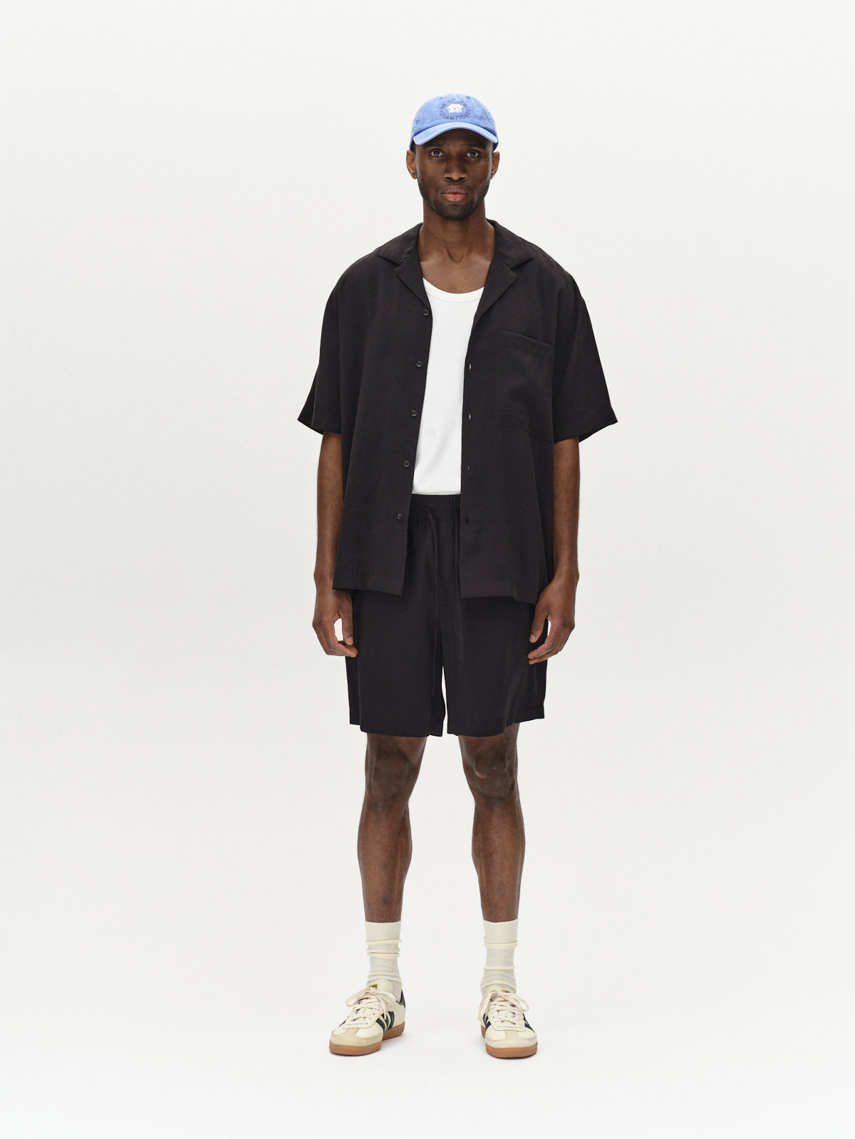 A man stands against a plain light background, wearing a light blue cap and a white tank top under a Gabba Jens Over Resort Yari SS - Meteorite oversized black open shirt with a statement chest pocket. He pairs it with black shorts, cream socks, and sneakers with dark accents.