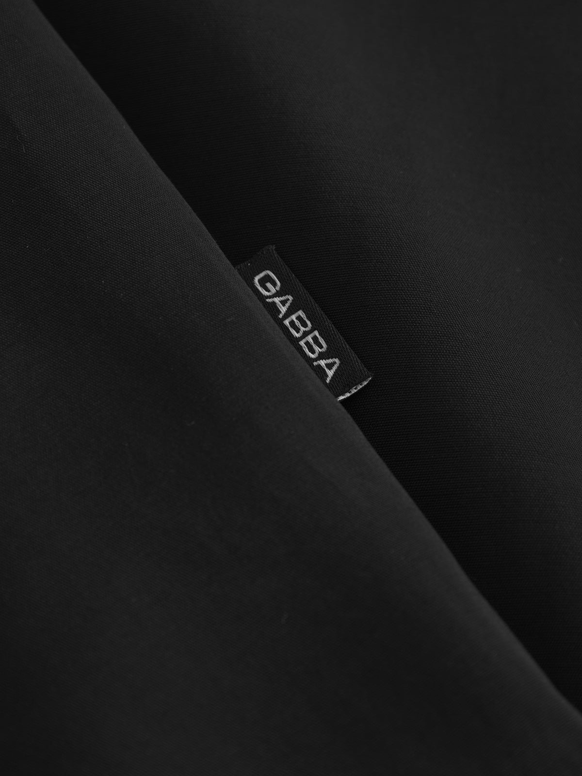 A close-up of the black fabric from the Jens Over Resort Yari SS - Meteorite by Gabba shows a small black clothing label with GABBA in white, partially tucked into a smooth, slightly reflective surface. Its understated elegance makes it ideal for any premium wardrobe.