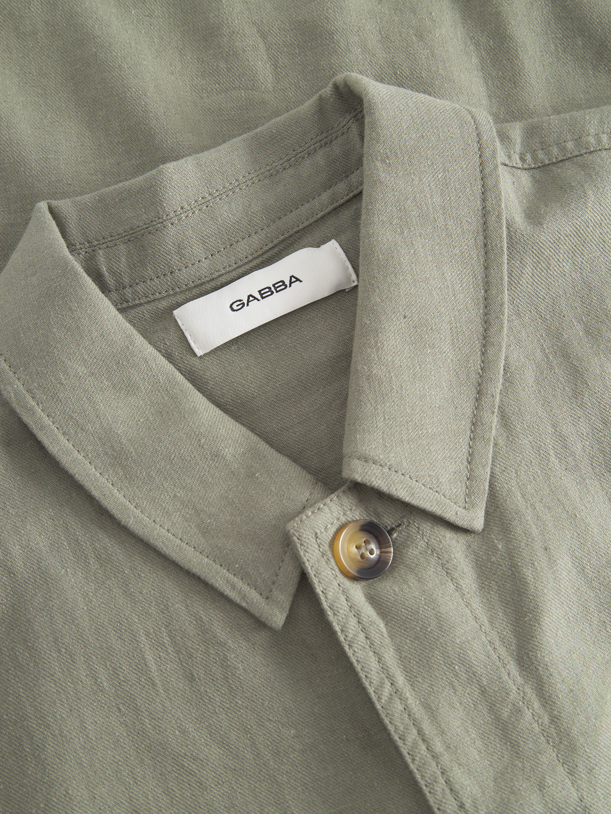 A close-up of the Peter Lino light green overshirt highlights the top button and collar, featuring a white label reading Gabba. The linen/cotton blend fabric looks soft and lightweight, making it an ideal spring companion.