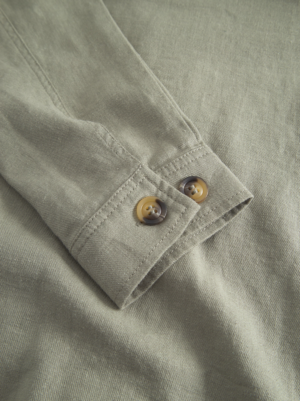 Close-up of the Peter Lino - Light Green linen/cotton fabric by Gabba with a sleeve detail showing two brown buttons. The texture reveals a casual, lightweight material, perfect as a spring companion or light overshirt for effortless style.