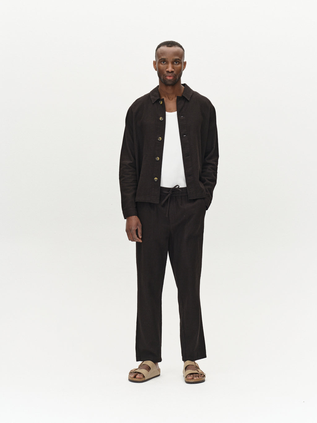 A man faces forward against a white background, wearing the Gabba Peter Lino - Meteorite overshirt and matching pants over a white shirt, paired with beige sandals—an ideal spring look. One hand is in his pocket.
