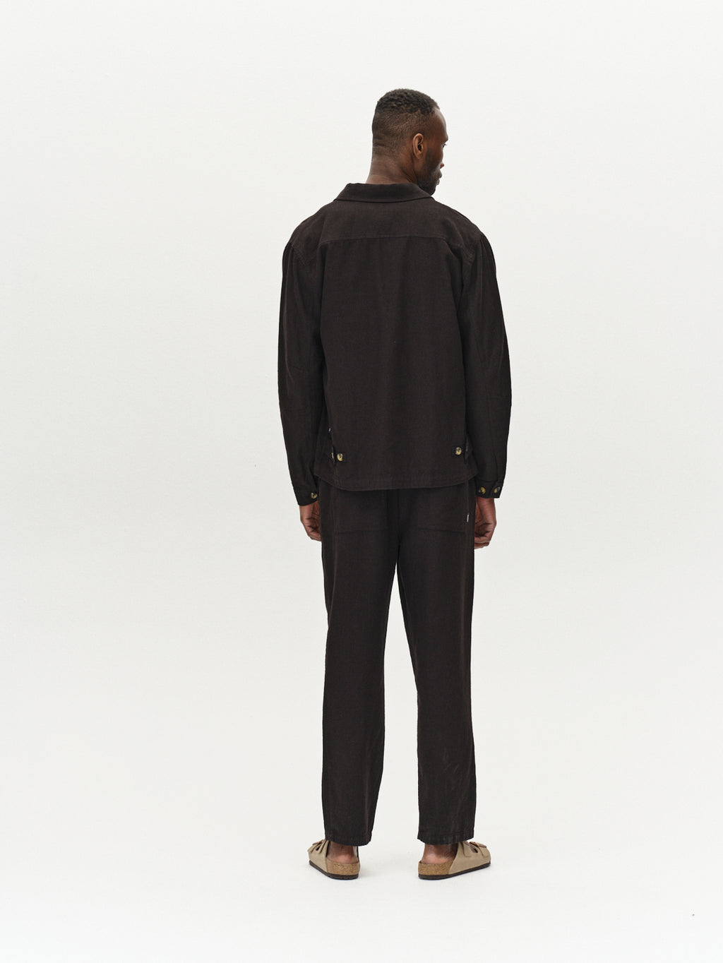 A person faces away from the camera, wearing Gabba’s Peter Lino - Meteorite loose black linen/cotton overshirt and matching pants, paired with brown slip-on sandals against a plain white background.