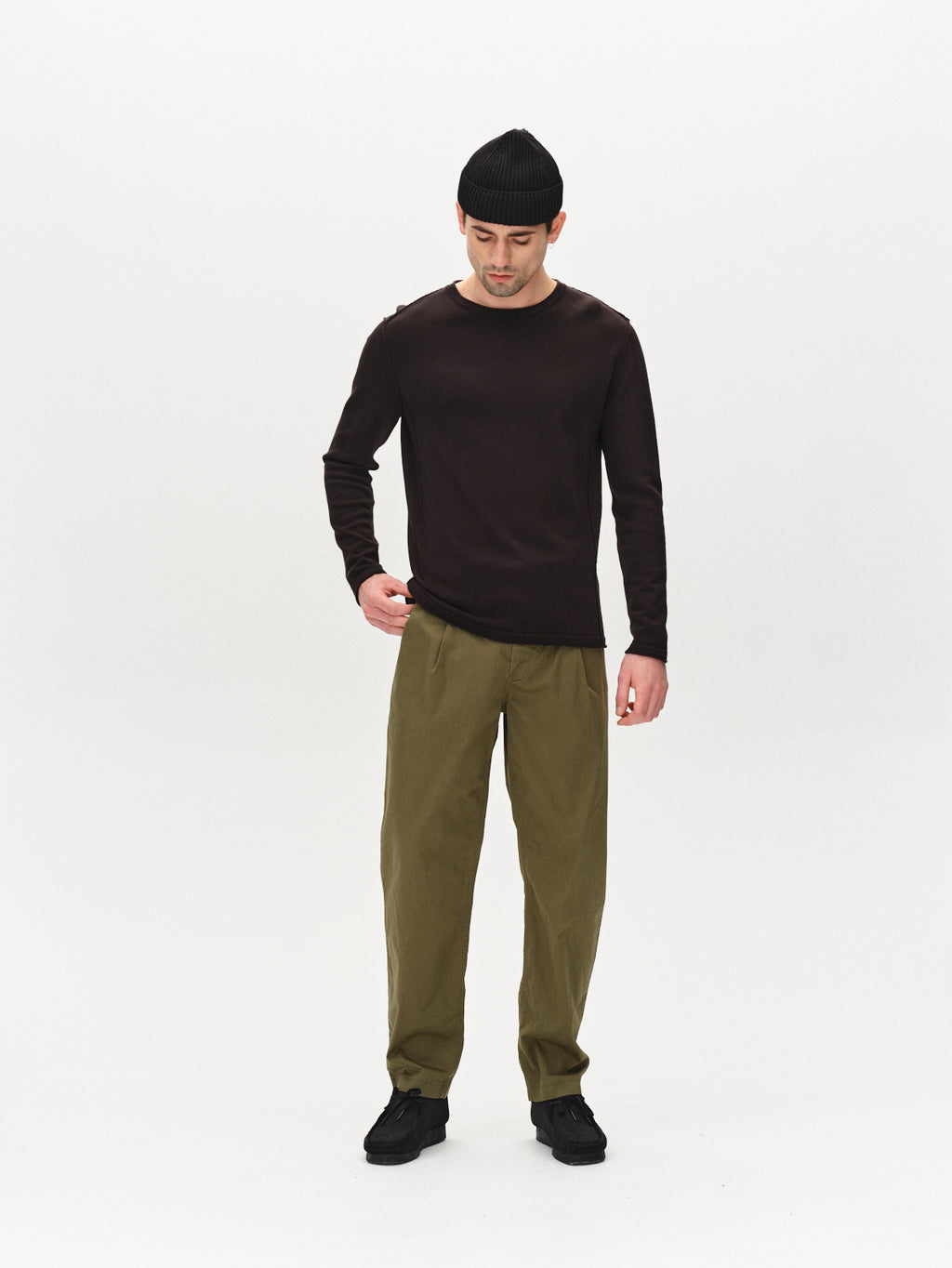 A man stands against a plain white background wearing the Gabba Bela Fix LS - Meteorite long-sleeve shirt, olive green pants, black knit beanie, and black shoes, looking down with one hand by his side and the other relaxed.