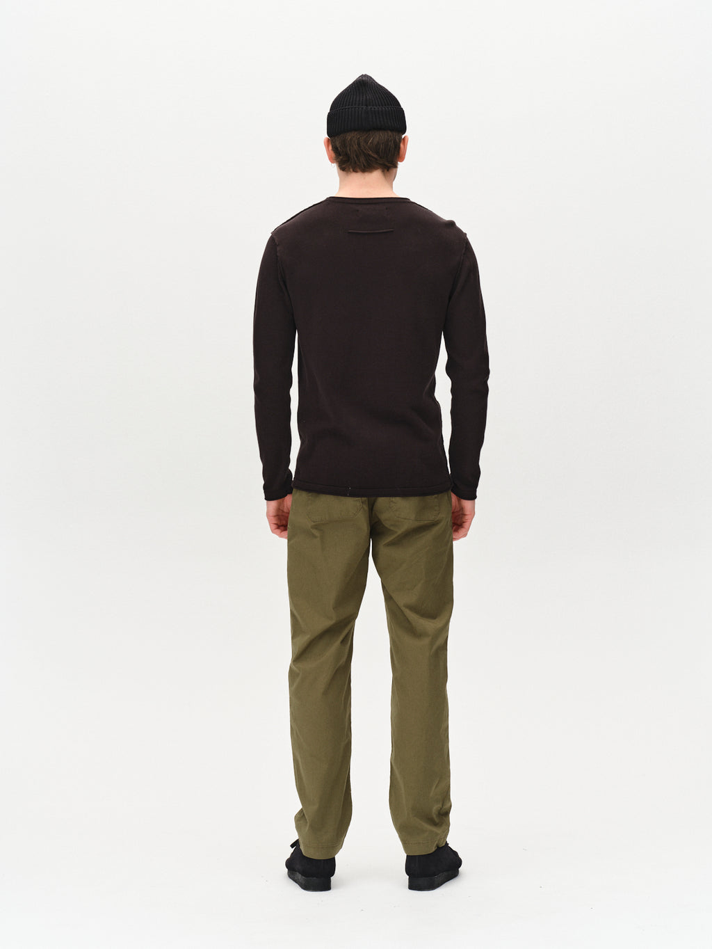 A person faces away from the camera, wearing Gabbas Bela Fix LS - Meteorite long-sleeve shirt in dark brown, paired with a black knit beanie, olive green pants, and black shoes against a plain white background for a stylish look.