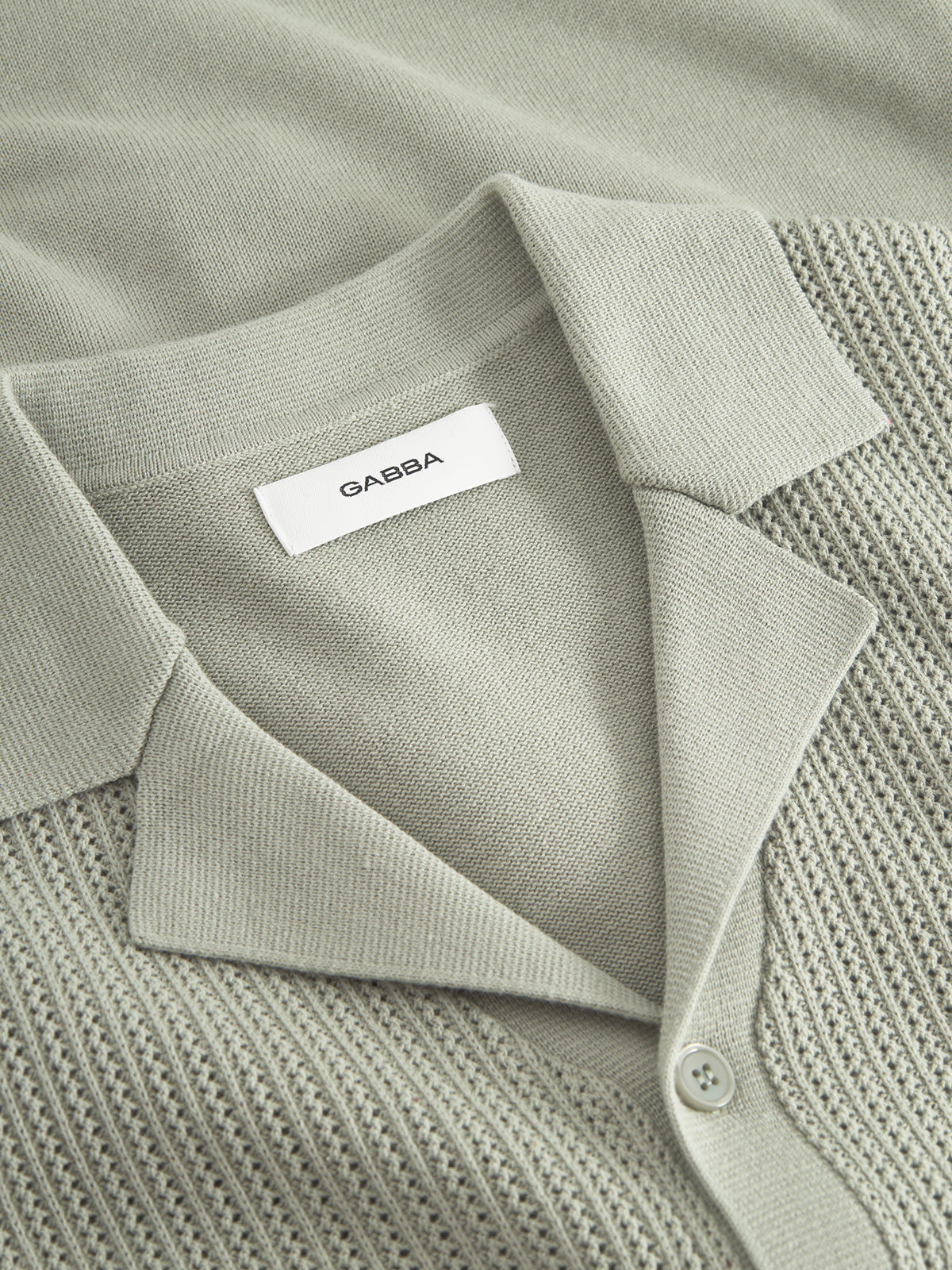 Close-up of a light gray textured knit shirt resembling a premium cardigan, featuring a collar and buttons. The inside label reads Gabba. The fabric is 100% cotton with a ribbed pattern near the bottom. Product: Geno Resort SS - Light Green by Gabba.