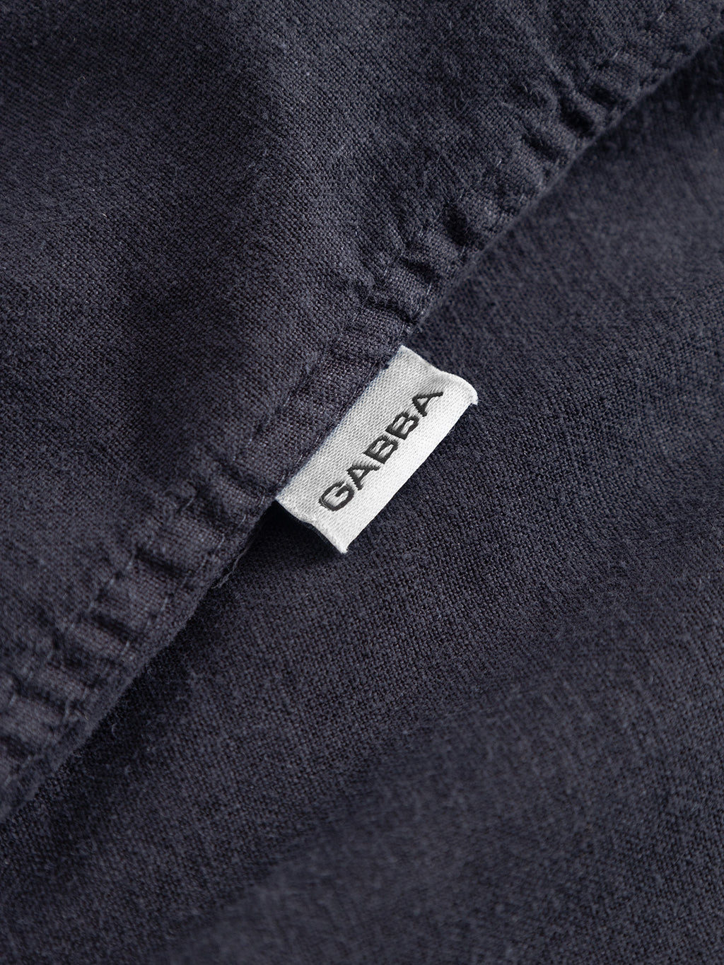 A close-up of the Sven Reg Lino SS - Night Sky shirt by Gabba, showing its dark garment-dyed fabric and a white tag with the Gabba brand name in black letters sewn into the seam.
