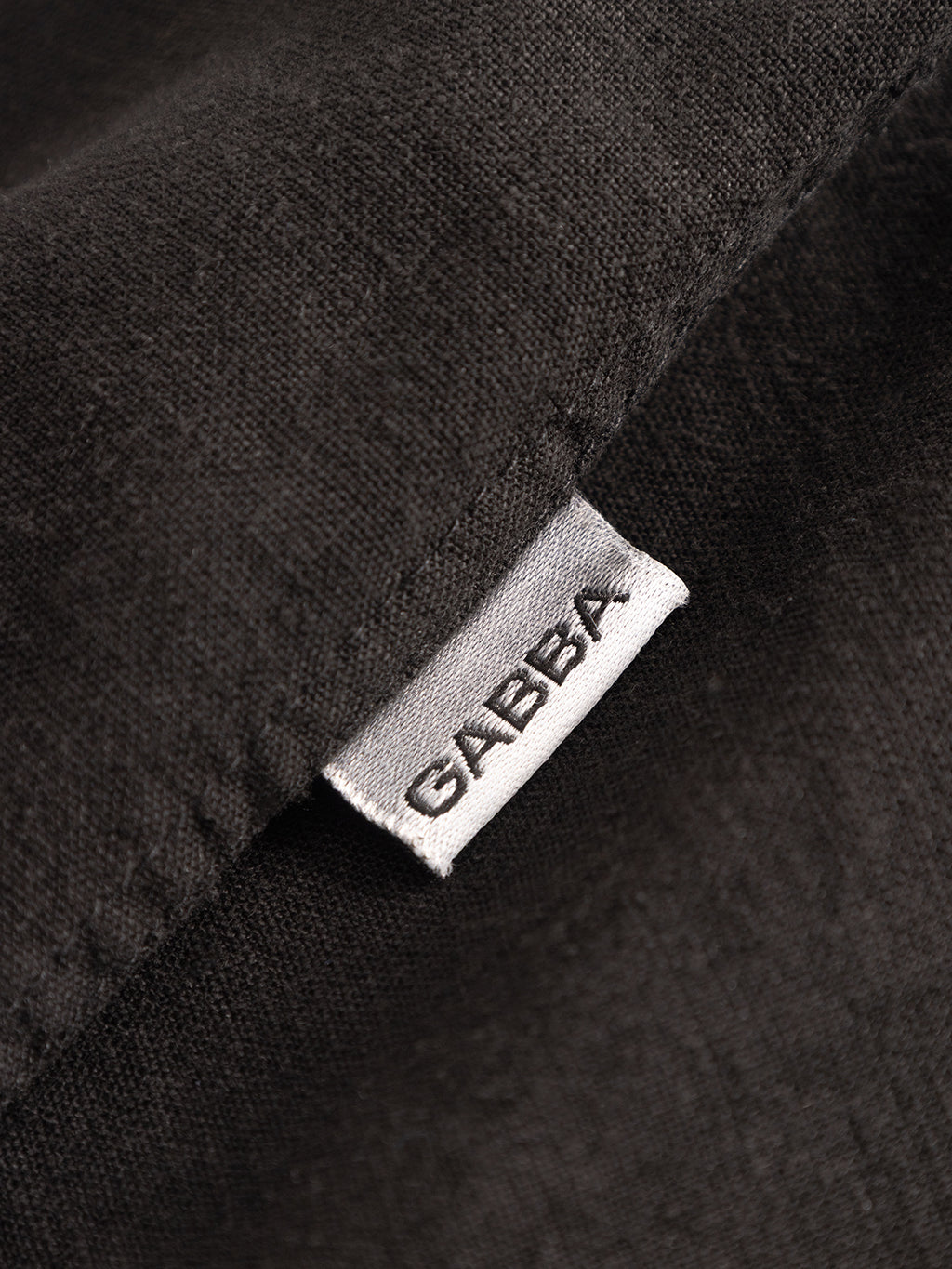 A close-up of a white fabric tag reading Gabba in black letters, sewn onto the textured dark linen of the Sven Reg Lino LS - Meteorite premium shirt.