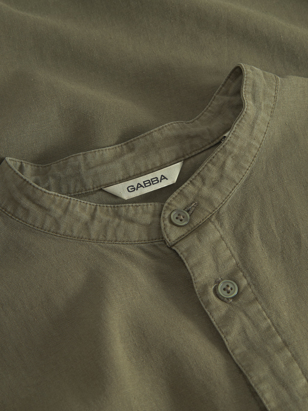 Close-up of the Gabba Sven Reg Lino Mand LS, a collarless shirt in olive green with two neck buttons fastened. Made from a lightweight, slightly textured linen-viscose blend; inner label displays Gabba.