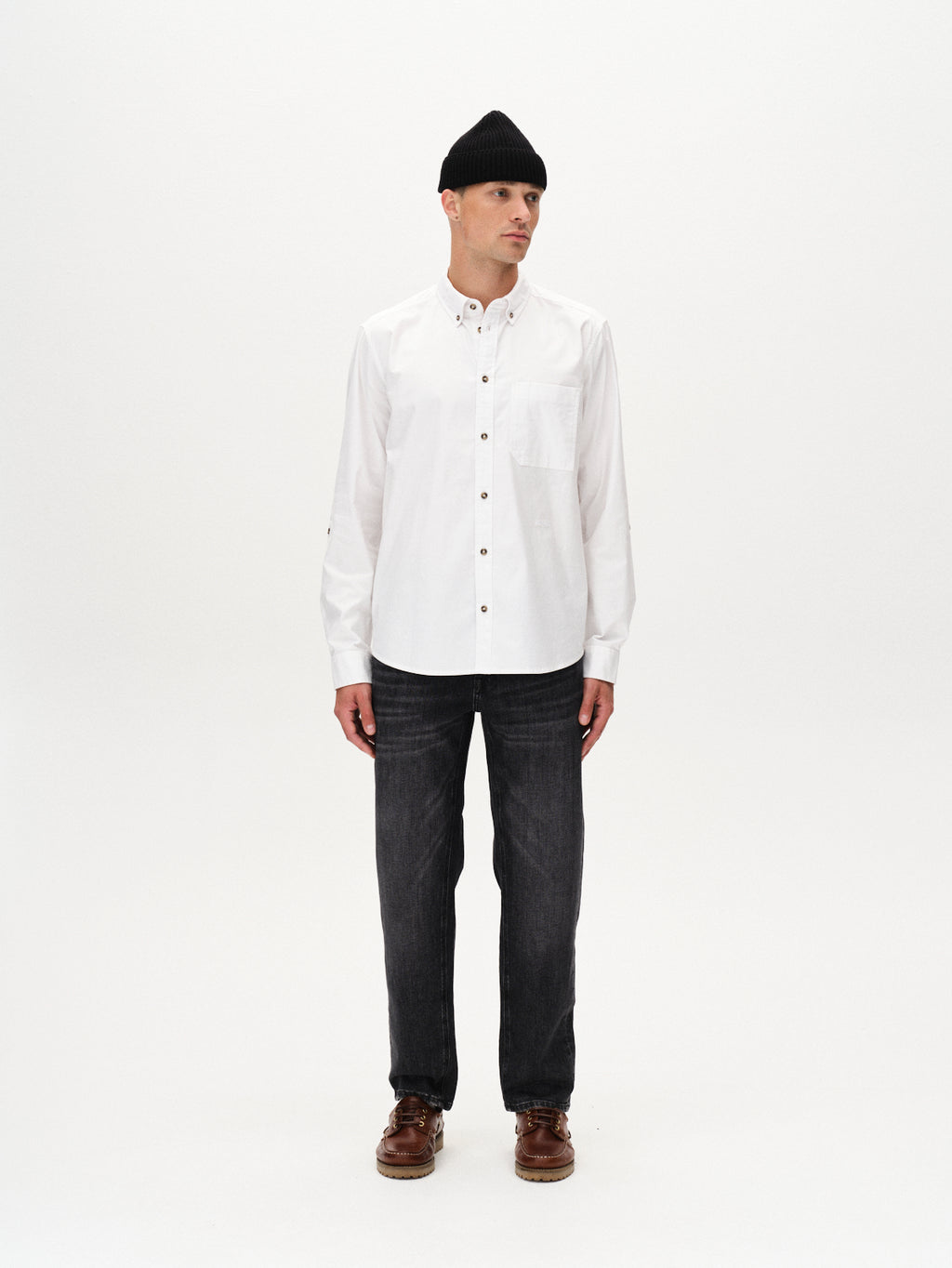 A man faces forward, wearing a black beanie, a white button-up shirt, Gabba Max F1011 - Black Denim slim fit jeans with Wiser Wash technology, and brown shoes, set against a plain white background.