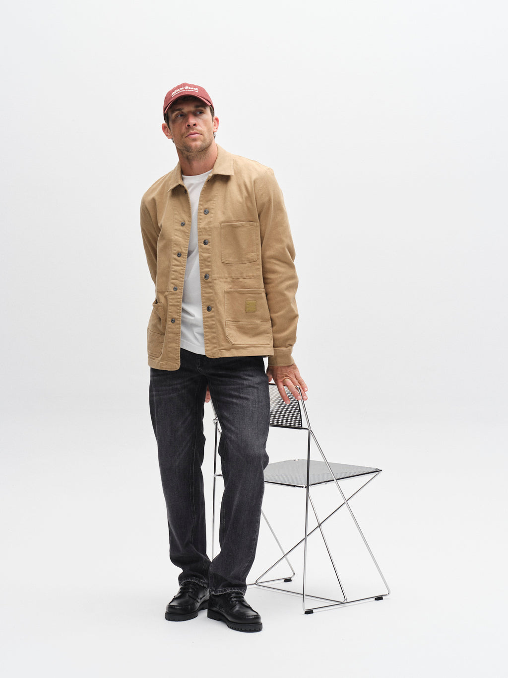A man in a red cap, beige jacket, white shirt, and black shoes wears Gabba’s GABmax F1011 - Black Denim jeans, standing by a modern folding chair against a neutral white backdrop.
