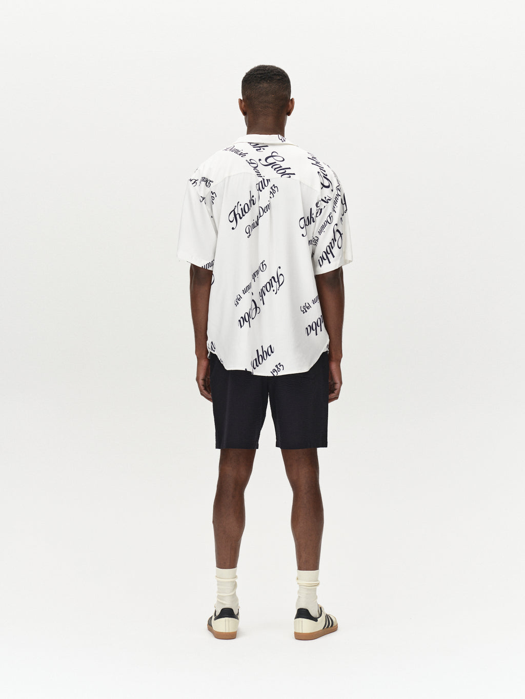 A man stands facing away in a Gabba Jens Over Resort Kiosk SS shirt with a navy pattern, paired with black shorts, cream socks, and sandals against a plain white background. The premium shirt is made from 100% Viscose.