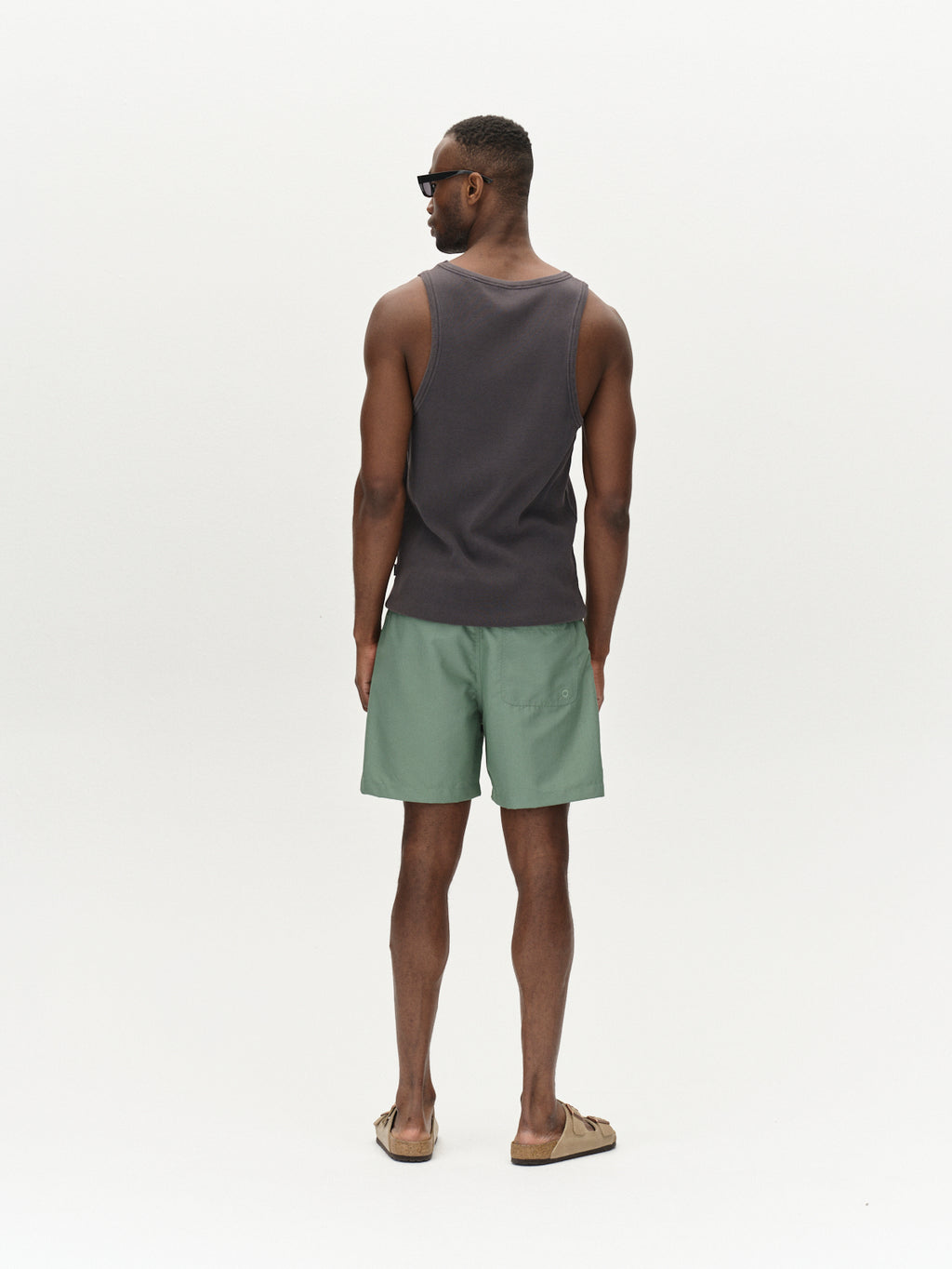 A man faces away from the camera, wearing Gabbas Jackson Plain - Green Sea swim shorts with an elastic waistband and side pockets, a dark sleeveless shirt, sandals, and sunglasses against a plain white background.