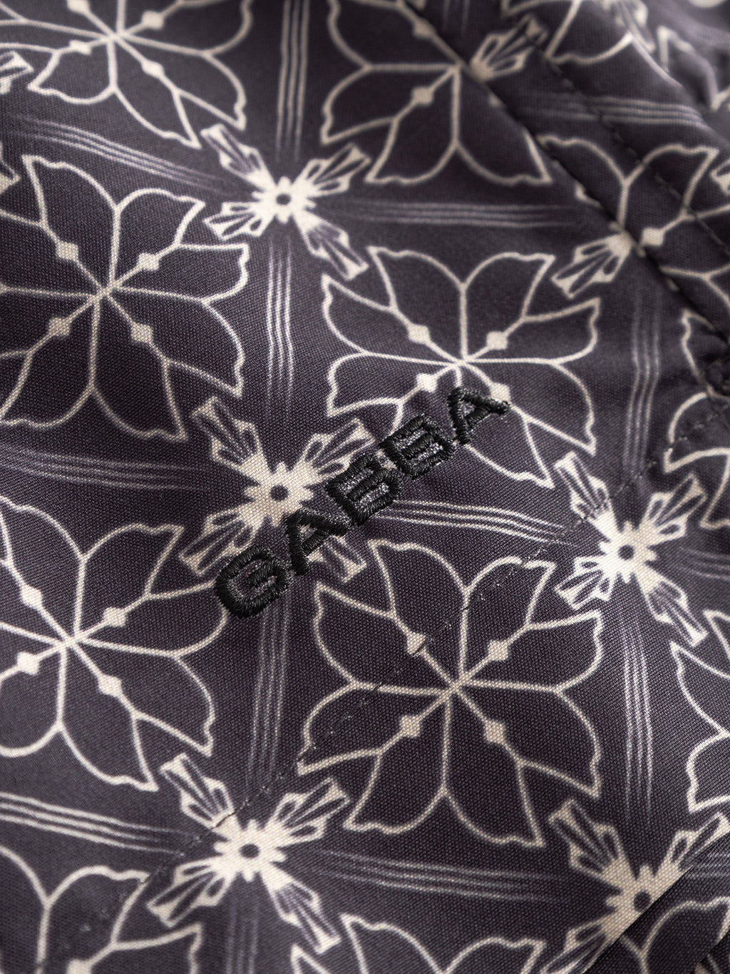 Close-up of Gabbas Jackson Print - Multi Pattern swim shorts fabric, featuring a dark base with a white geometric floral design and Gabba embroidered in black thread.