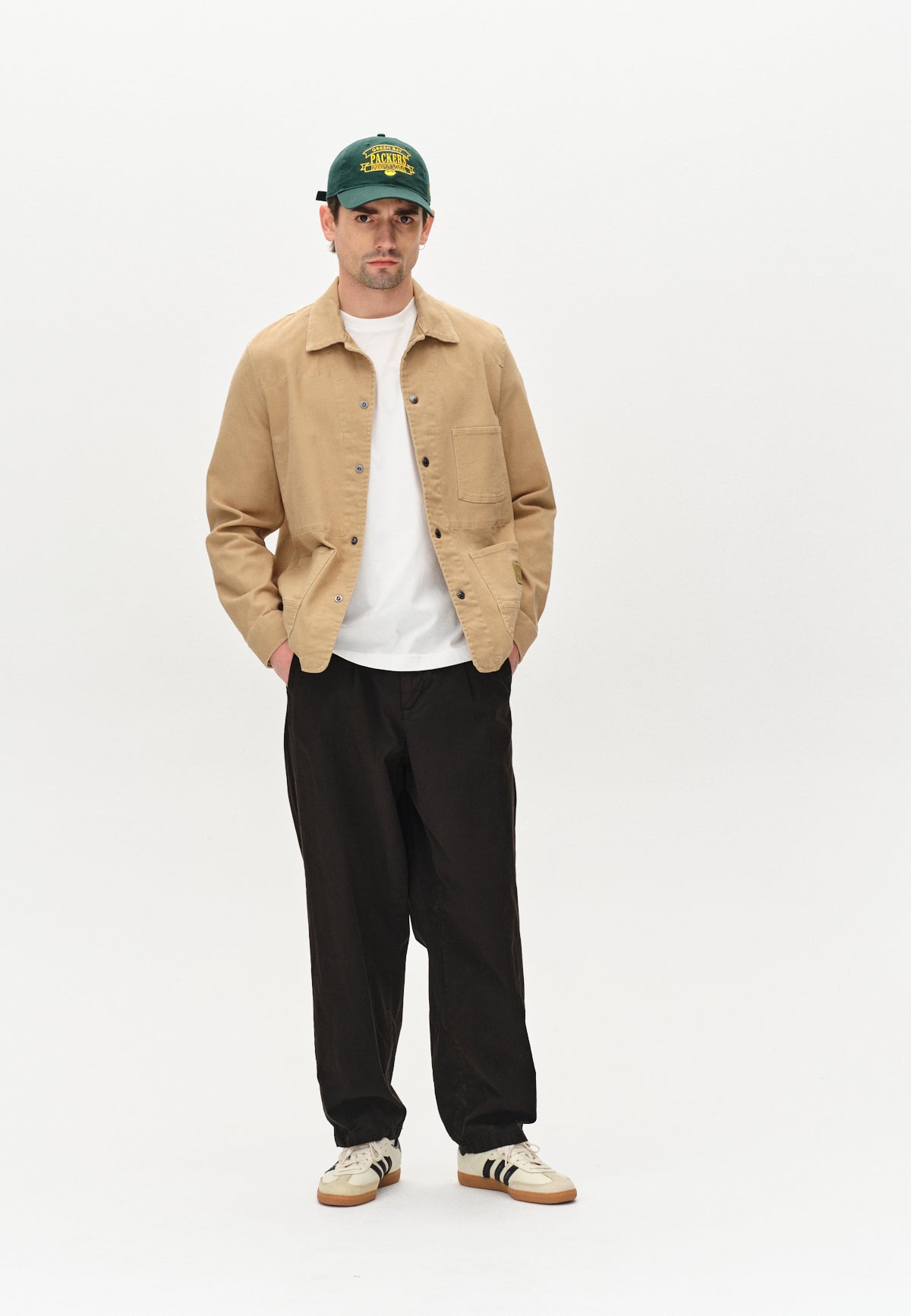 A person stands against a plain white background, hands in pockets, wearing Gabbas Beta Twill - Sand overshirt over a white tee, black pants, white sneakers with black stripes, and a green cap.