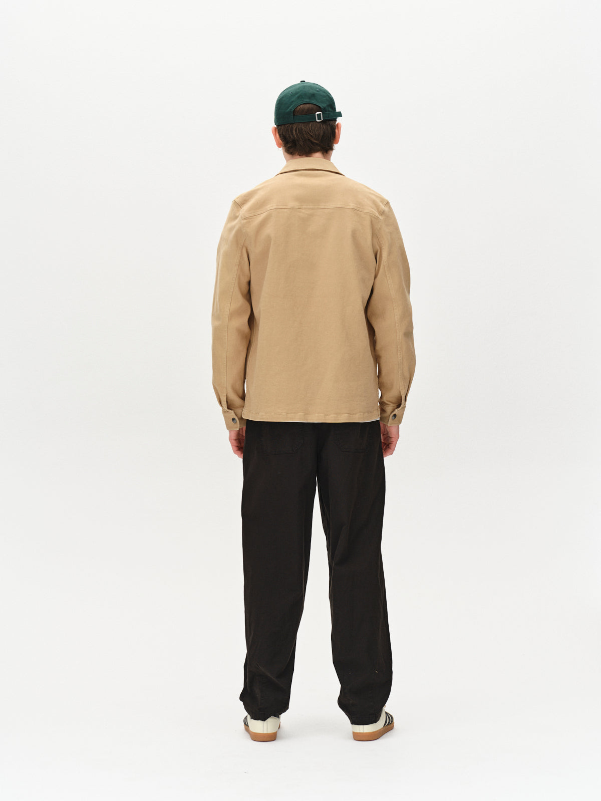 A person stands facing away from the camera, wearing Gabba Beta Twill - Sand overshirt, a green baseball cap, black pants, and white sneakers against a plain white background.