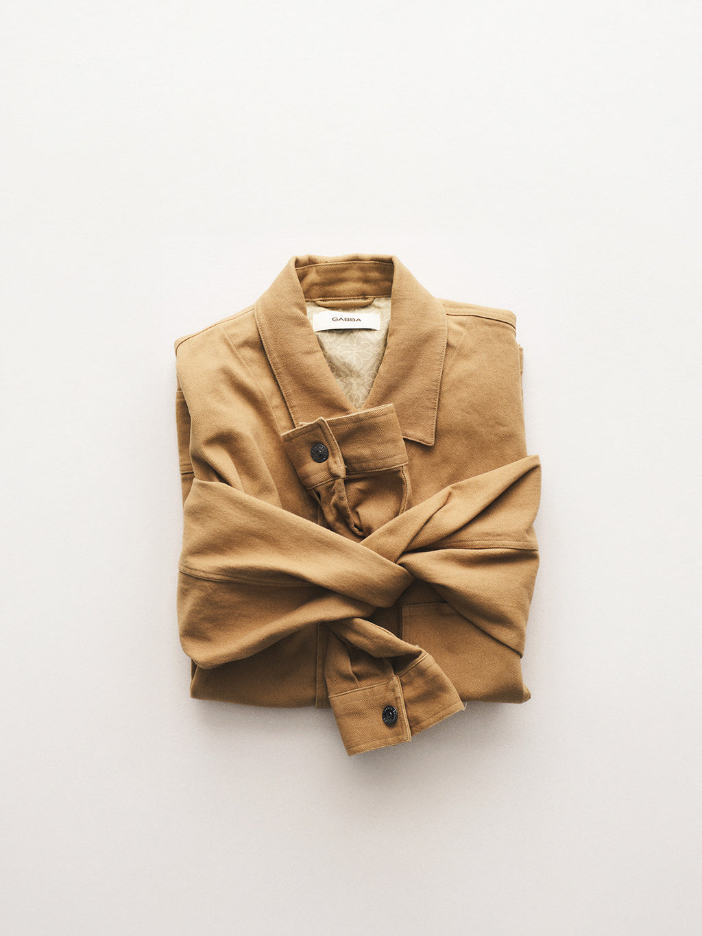 The Gabba GABbeta Twill - Sand collared overshirt, garment dyed for a refined workwear look, is neatly folded with crossed sleeves on a plain light background.