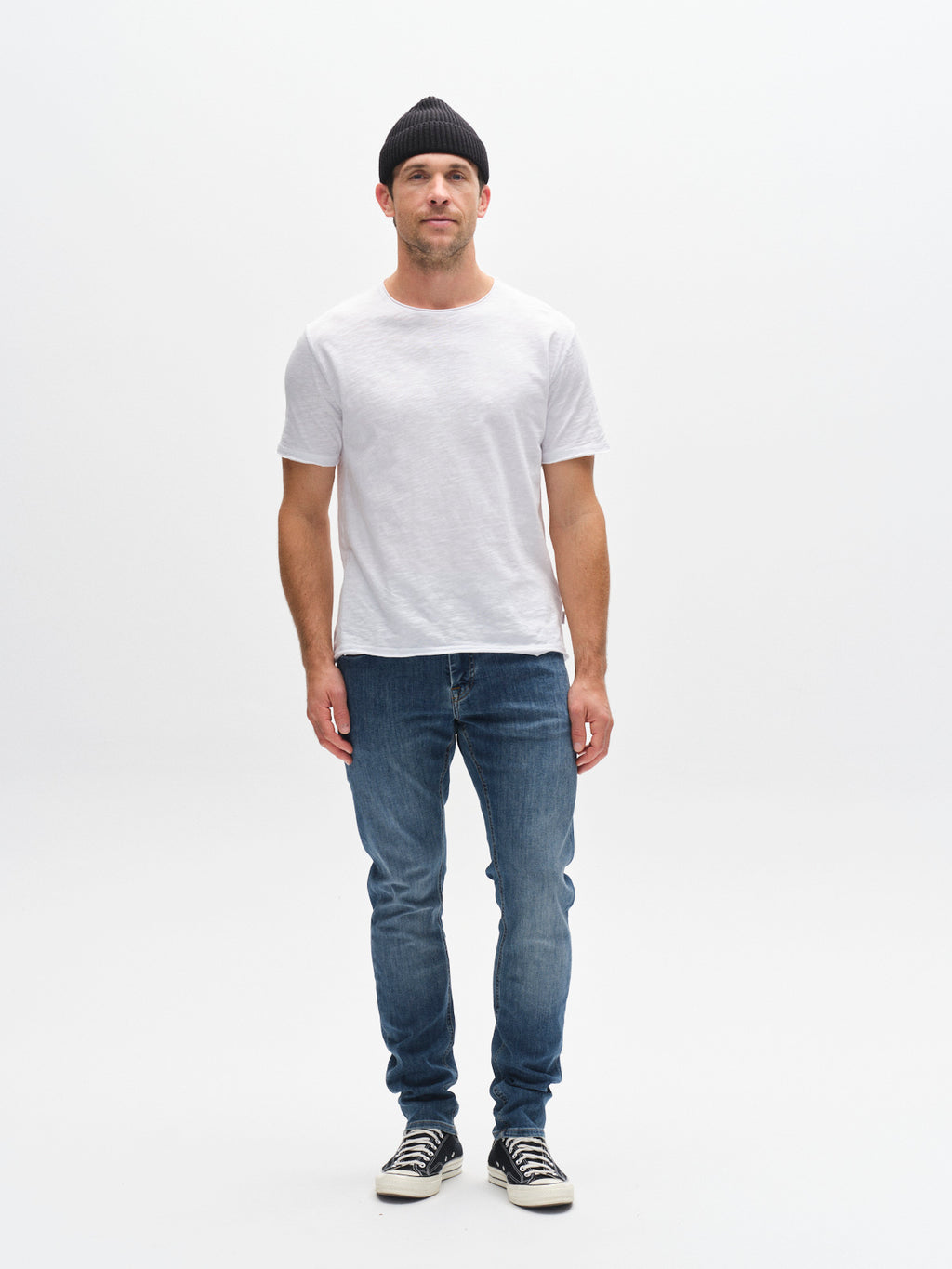 A man stands facing forward against a white background, wearing a black beanie, plain white t-shirt, Gabba GABjones Vanew Mid Blue Denim jeans with a slim fit and super-stretch fabric, and black-and-white sneakers.