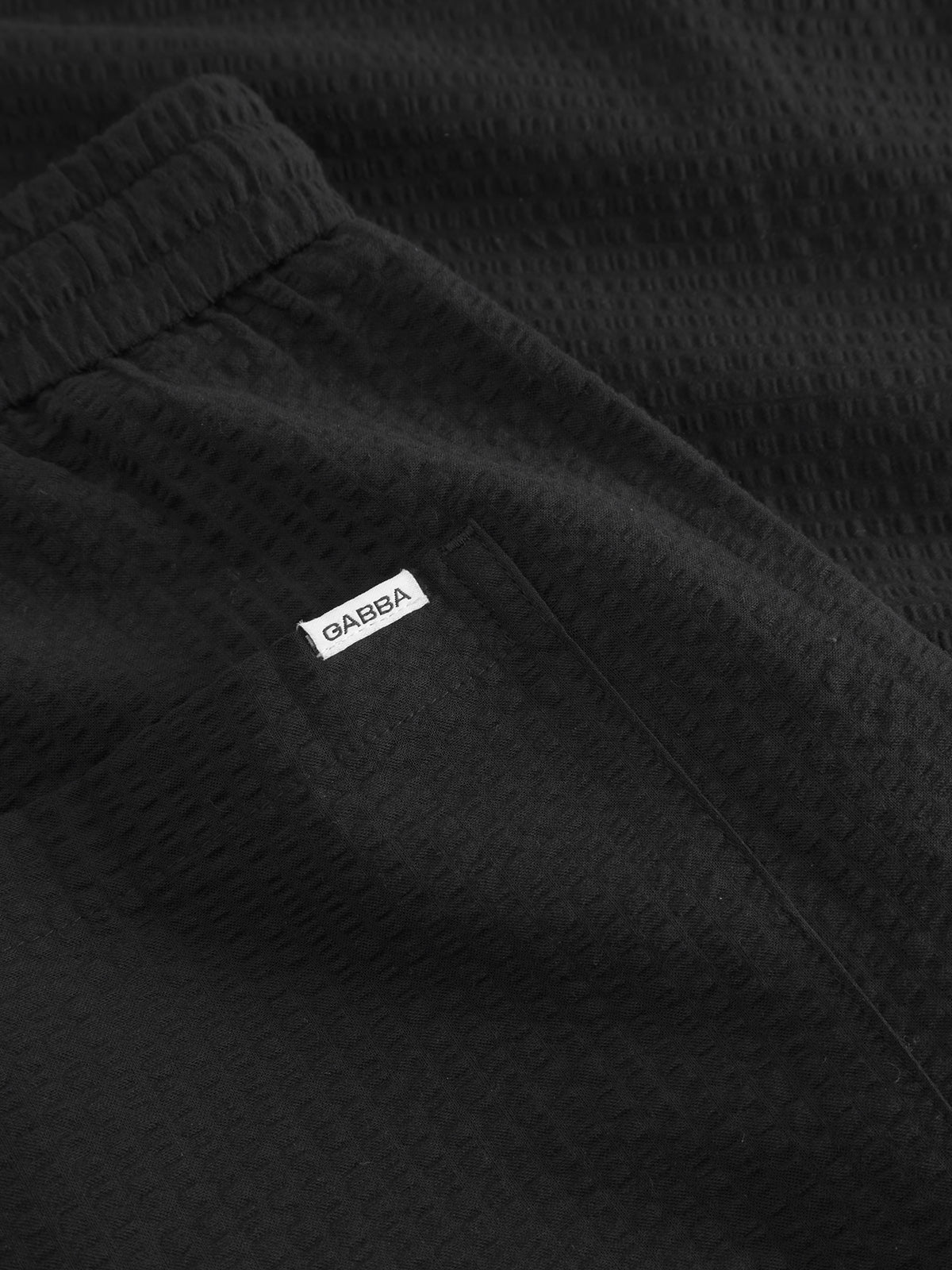 Close-up of textured black Tommi Seer pants by Gabba, made from 100% cotton with an elastic waistband and a small pocket featuring a white GARBA label. The subtle grid-like pattern resembles seersucker, offering a unique look.