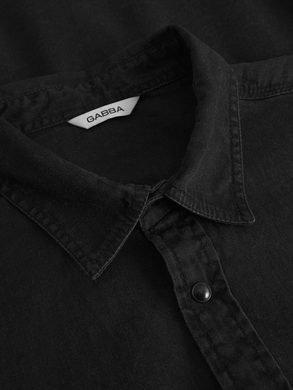 Close-up of the Gabba Sven Reg Denim LS shirt in black denim, made from 100% cotton with a pointed collar and snap button. A white Gabba label is visible inside the collar along with detailed fabric texture and stitching.