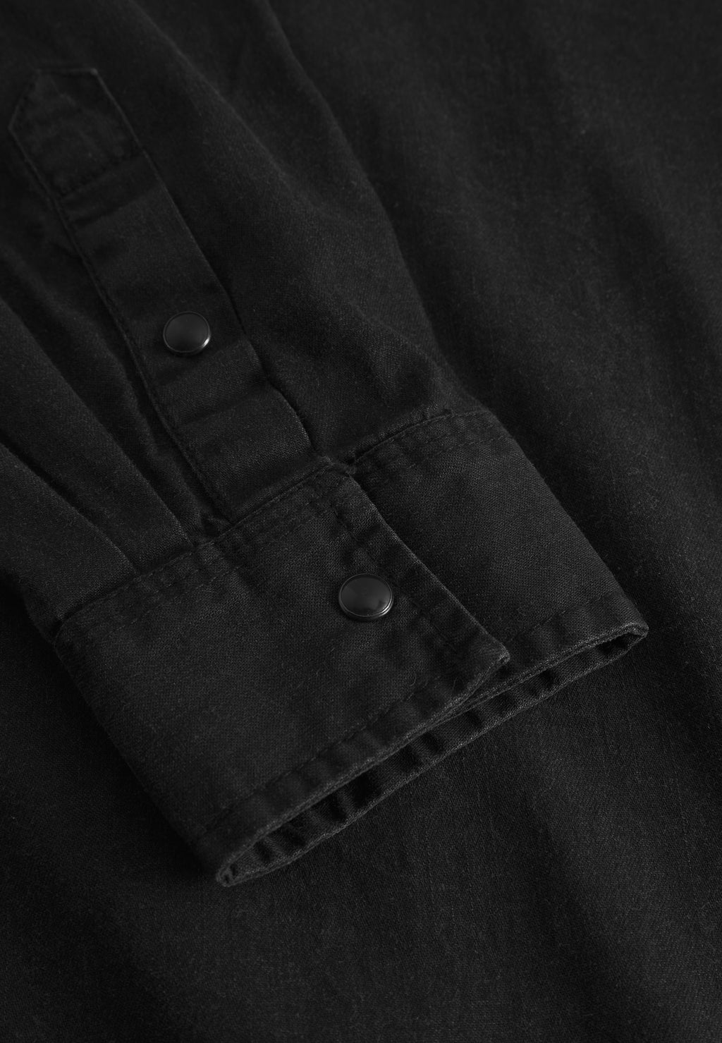 Close-up of the Sven Reg Denim LS - Black Denim shirt by Gabba, featuring a black denim sleeve with two black metal snap buttons on the cuff, visible stitching, and a slightly textured 100% cotton fabric.