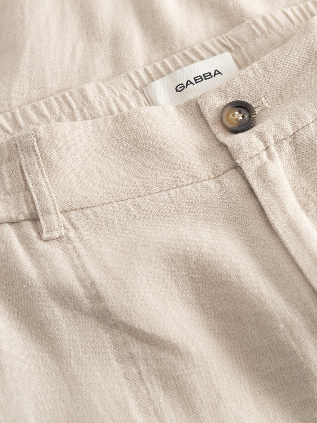 Close-up of GABBA's GABzeta Lino - Birch pants in light beige, showing the waistband, belt loop, brown button, and white Gabba label. The lightweight linen fabric looks soft with a slight texture.