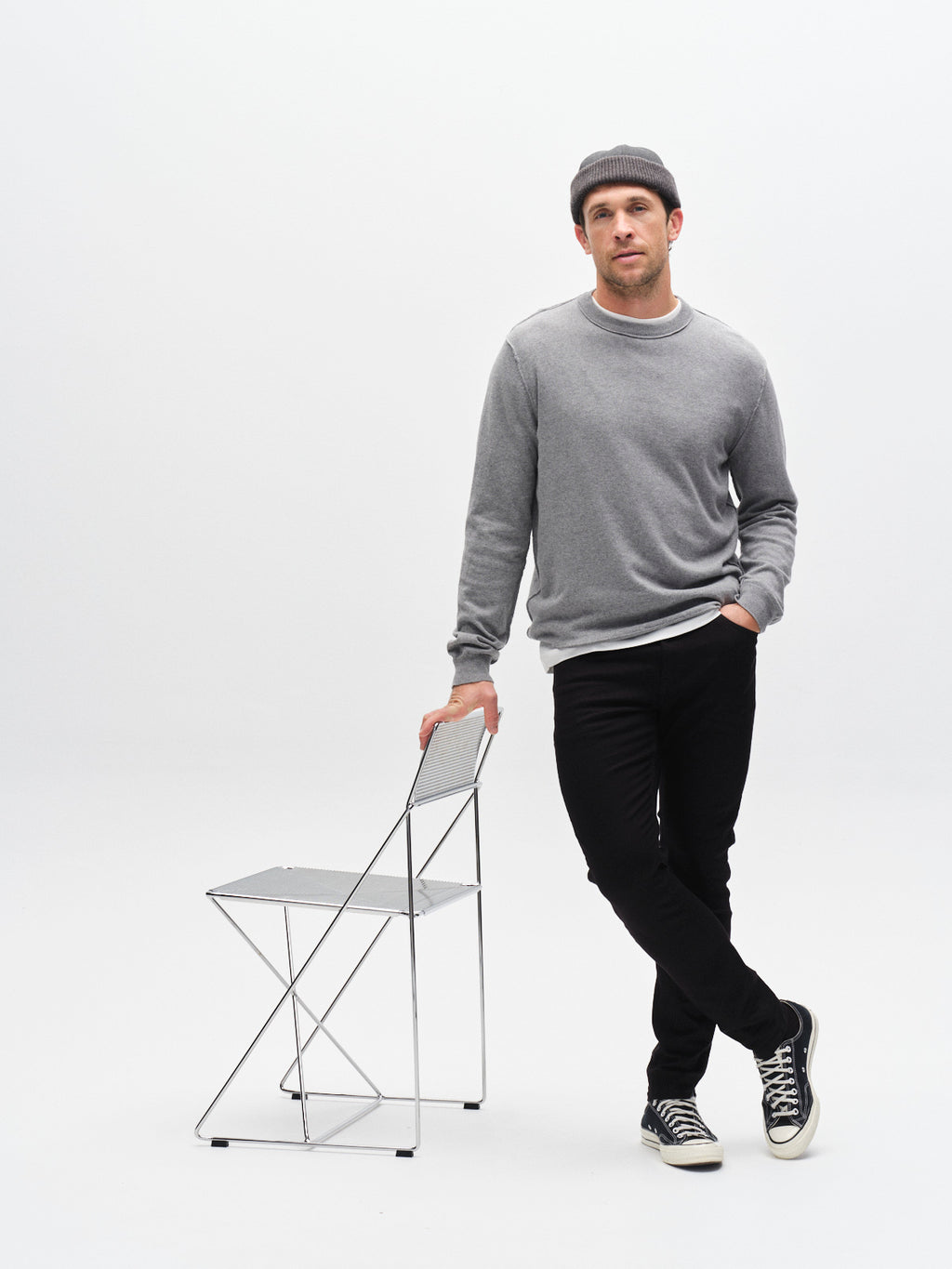 A man in a gray sweater, black sneakers, gray beanie, and Gabba’s GABjones Hold Black - Black Denim jeans leans casually against a metal folding chair in a minimal white studio setting.