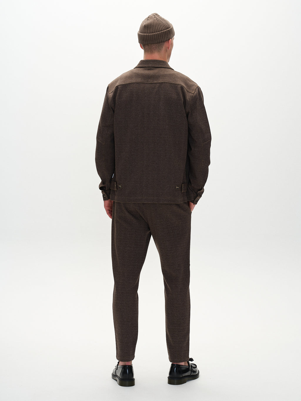 A person faces away, wearing a brown jacket, Gabbas Monza Tweed pants (Brown), and a knitted beanie that elevates their sophisticated style, complemented by black sandals against a plain, light-toned backdrop.