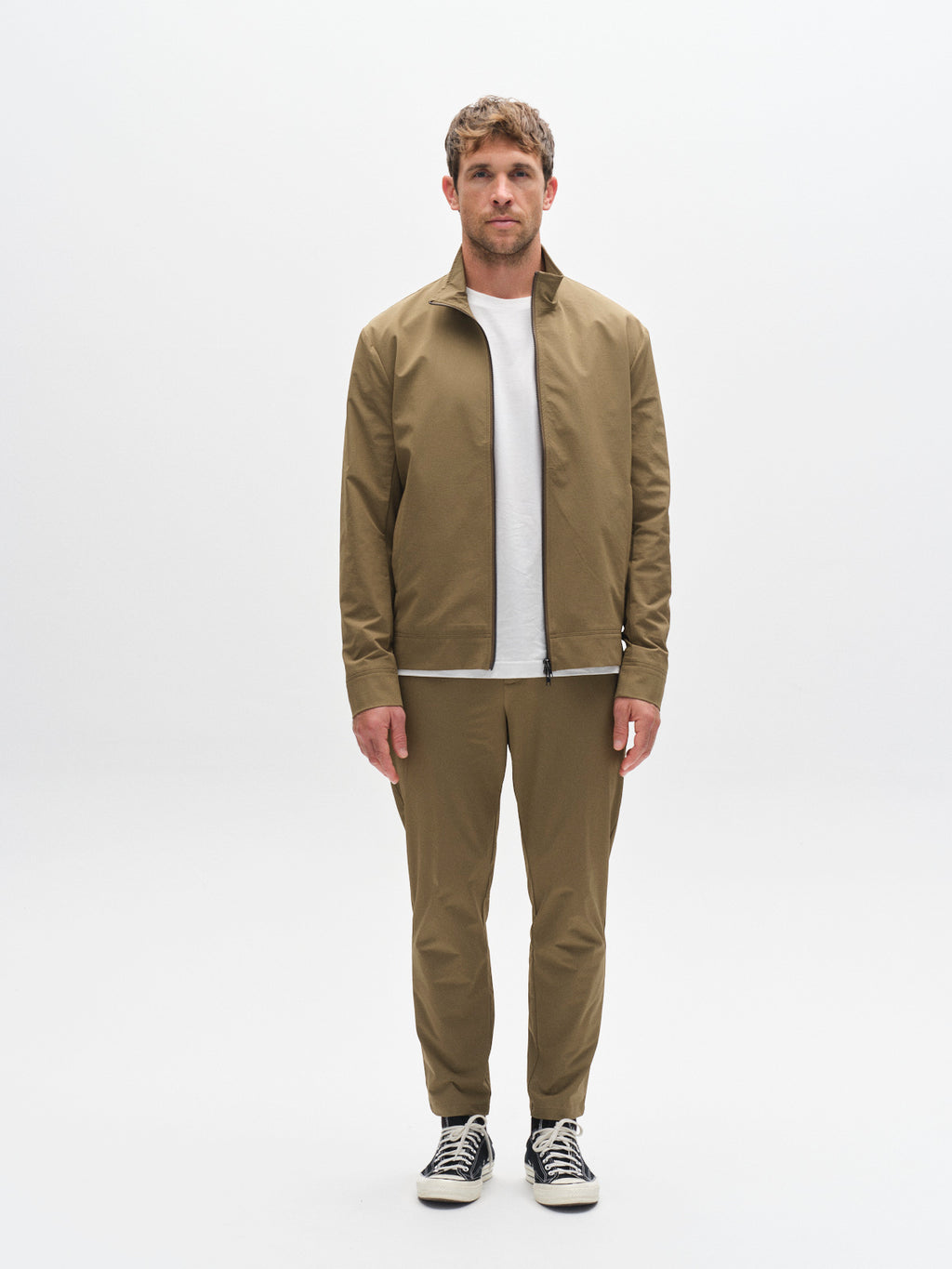 A man stands facing forward against a plain white background, wearing the Gabba GABnik Tech - Army jacket and matching pants, paired with a white t-shirt and black sneakers with white soles and laces.