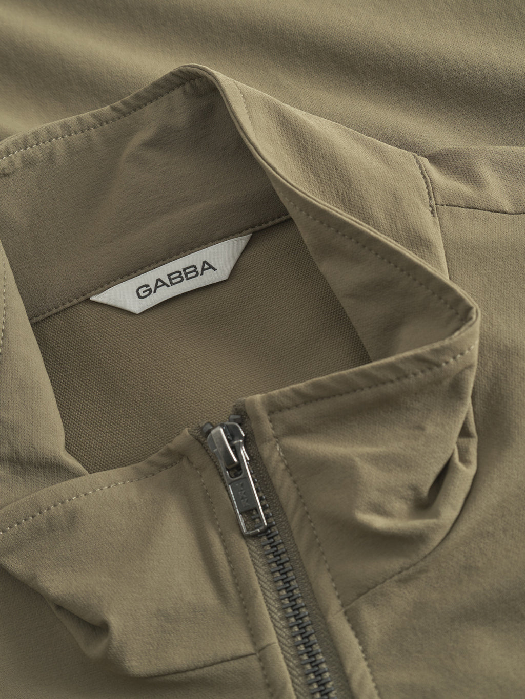 Close-up of the Gabba GABnik Tech - Army jacket in khaki, showcasing its modern design with a visible collar, smooth fabric, partially unzipped front, and a white Gabba label inside the neckline.