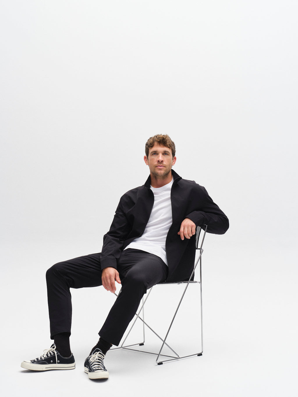 A man wearing the Gabba GABnik Tech jacket in black, paired with a white t-shirt, black pants, and black sneakers, sits on a minimalist metal chair against a plain white background, looking at the camera.