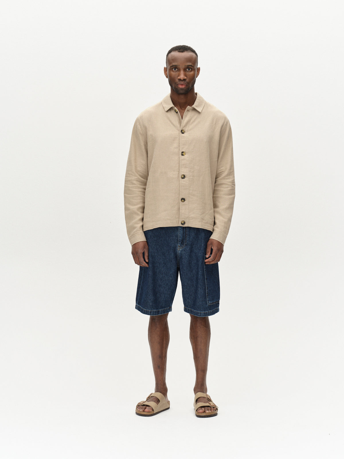 A person stands against a plain background, wearing a beige button-up shirt and Gabbas Ugo Cargo Fone shorts in Mid Blue Denim with a laid-back fit, paired with casual sandals. Their hands rest at their sides, capturing an effortless workwear vibe.