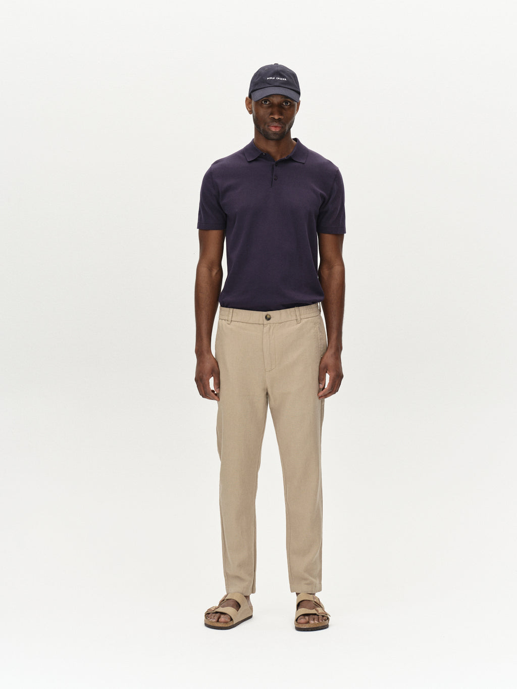 A man stands facing forward against a white background, arms relaxed, wearing Gabba’s Monza Lino Cropped - Sand Plaza pants with a navy cap, dark polo, and brown sandals, showcasing a naturally relaxed silhouette.