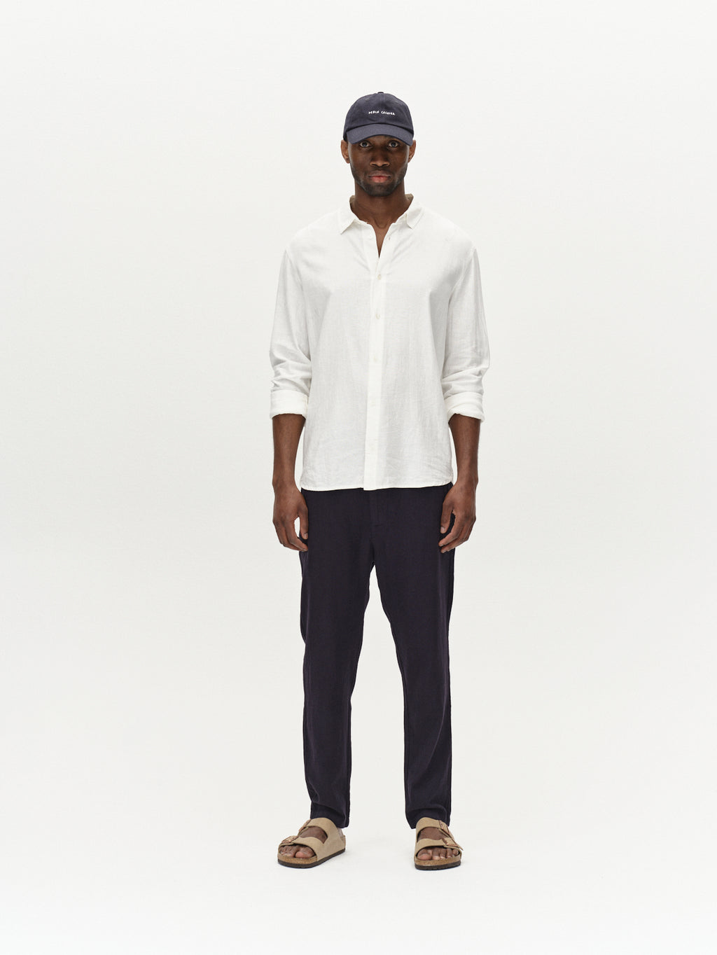 A man faces forward against a plain white background, wearing a navy cap, a white long-sleeve shirt, beige sandals, and Gabba Monza Lino Cropped pants in Night Sky with an elastic waist for comfort.