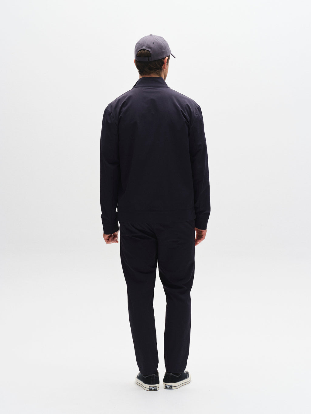 A person wearing a dark jacket, Gabba GABmonza Tech - Navy tech fabric pants with a relaxed straight fit, and a cap stands facing away, looking at a white background.