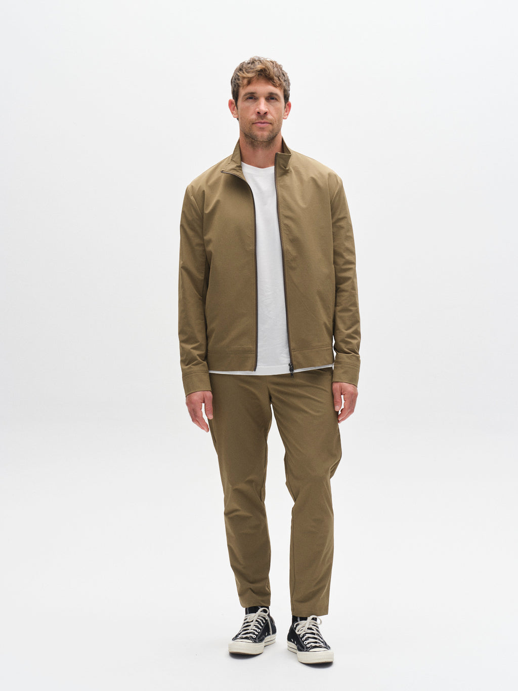 A man faces forward against a plain white background, wearing the Gabba GABmonza Tech - Army jacket, relaxed straight fit tech fabric pants, a white T-shirt, and black low-top sneakers with white soles.