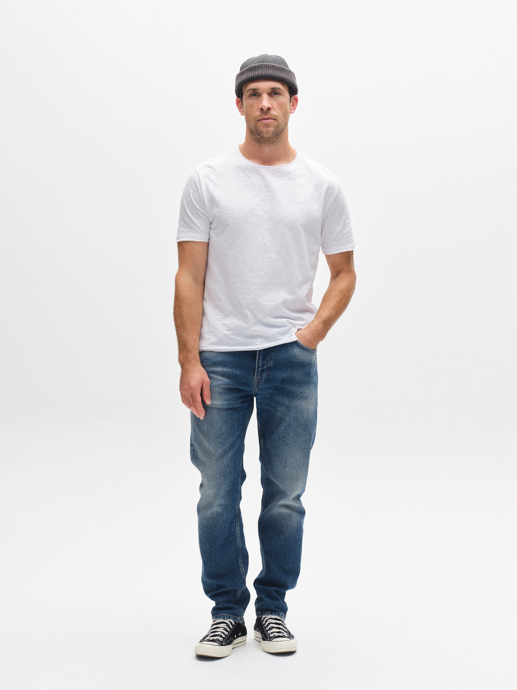 A man wearing the Gabba GABkostant New SS - White t-shirt, a gray beanie, blue jeans, and black sneakers stands against a plain white background, showcasing minimalist men’s fashion with one hand in his pocket.