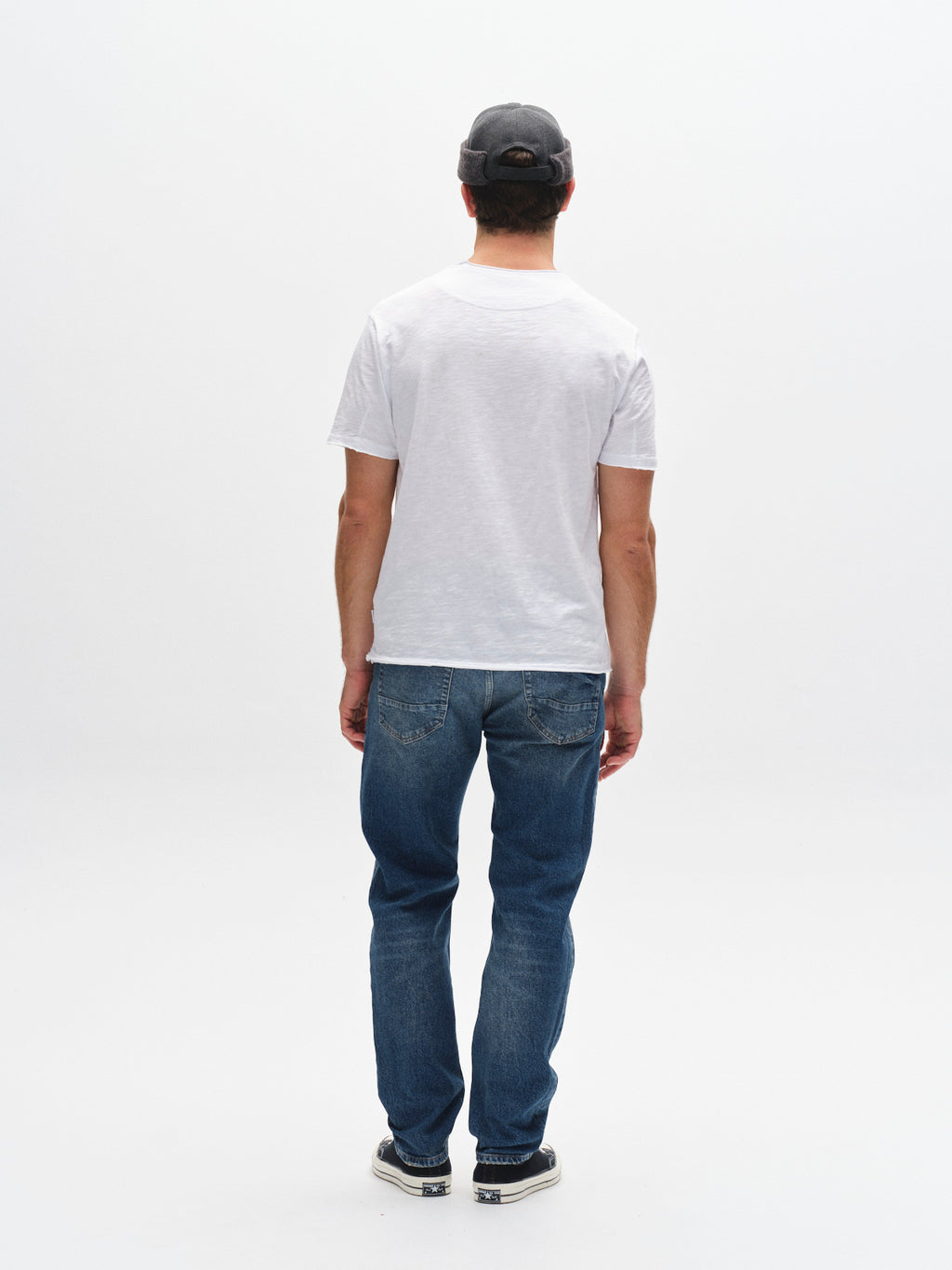 A man in a gray cap, blue jeans, and black shoes models the Gabba GABkostant New SS - White t-shirt, facing away from the camera against a plain white background to showcase minimalist men's fashion.
