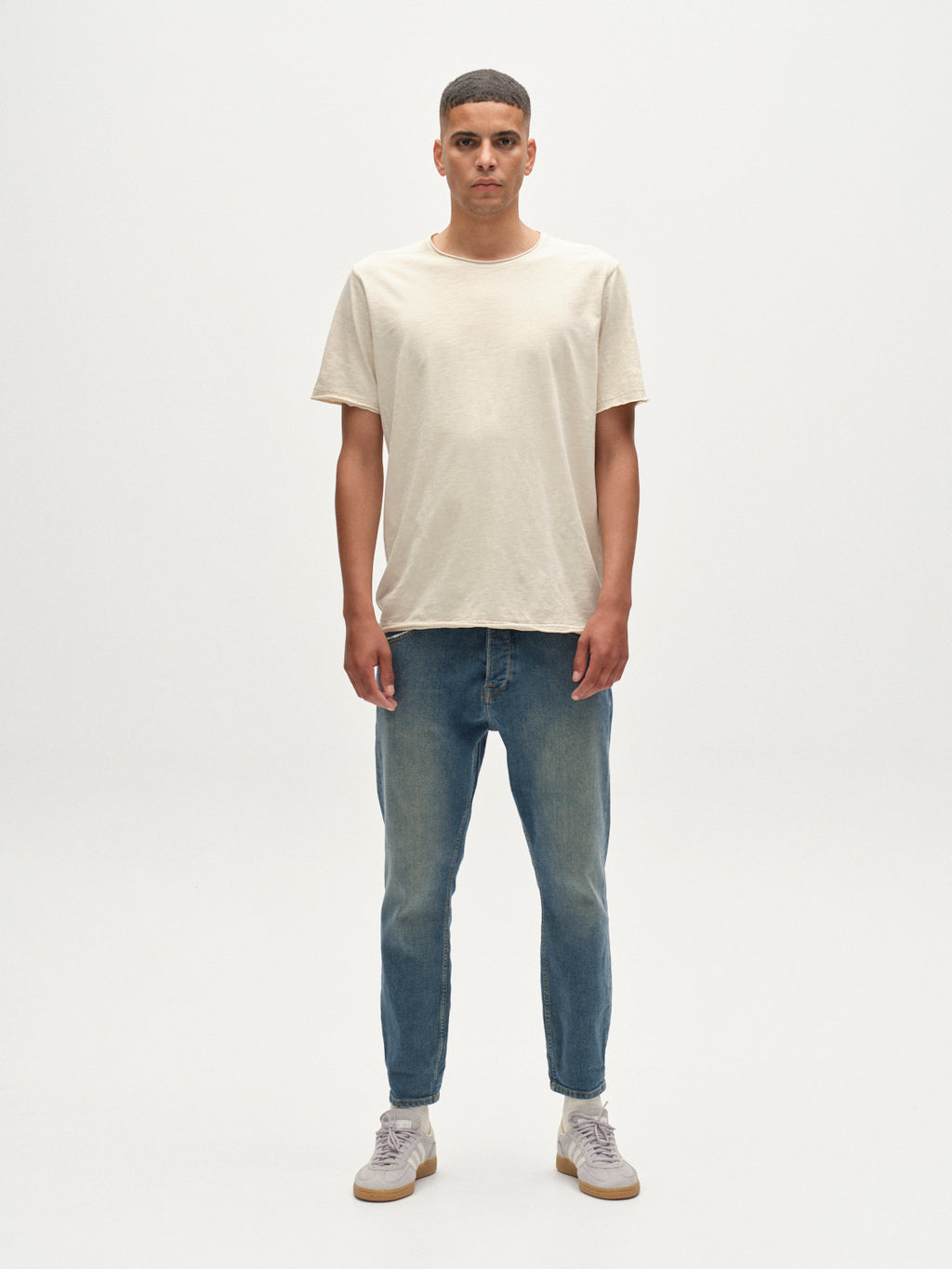 A man faces forward against a plain white background, wearing the Gabba Kostant New SS t-shirt in White Almond with a light slub texture, paired with blue jeans and light-colored sneakers.