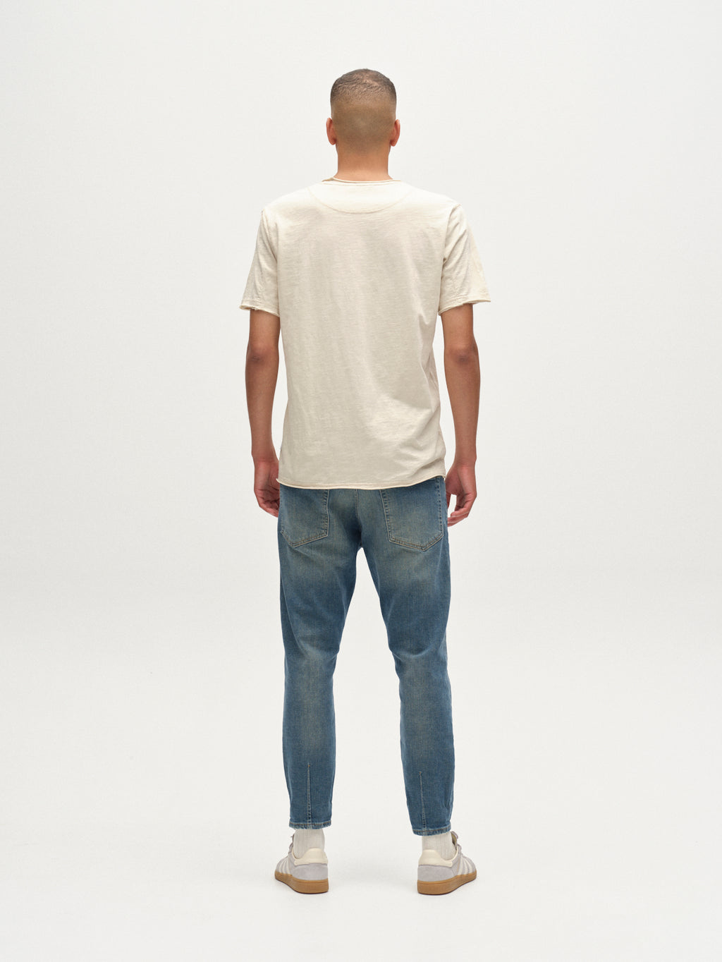 A person stands facing away from the camera wearing the Gabba Kostant New SS T-shirt in White Almond, paired with blue jeans and white sneakers, against a plain white background.