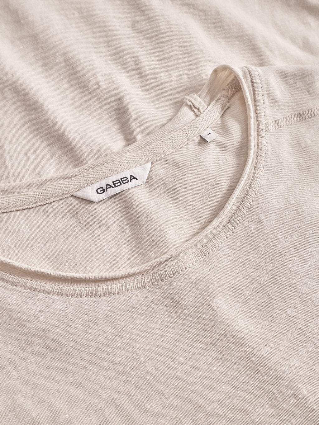 A close-up of the Gabba Kostant New SS in White Almond, a regular fit T-shirt with a round neckline, visible stitching, subtle light slub texture, and a white Gabba tag inside the collar.