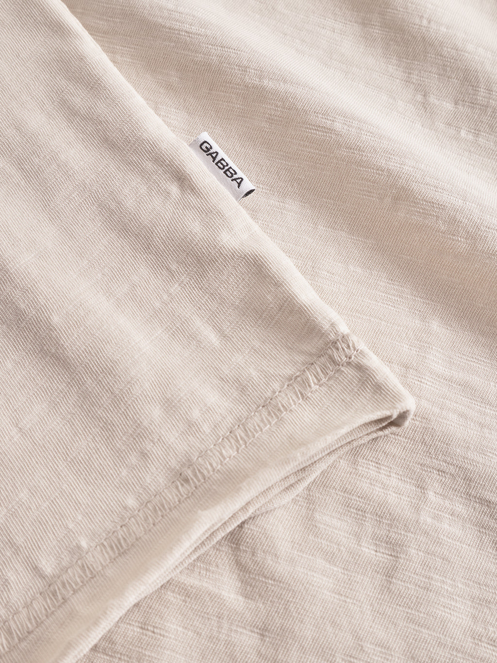 Close-up of Kostant New SS - White Almond by Gabba, featuring a light slub texture, regular fit, stitched hem, and a small white Gabba tag.