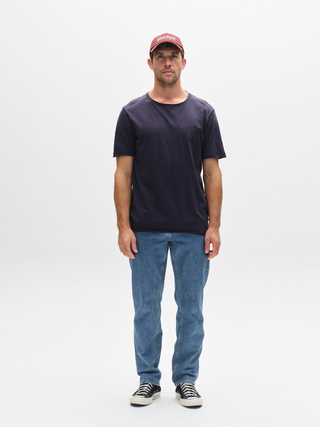 A man faces forward against a white background, wearing the Gabba GABkostant New SS navy slub t-shirt, blue jeans, black-and-white sneakers, and a red baseball cap.