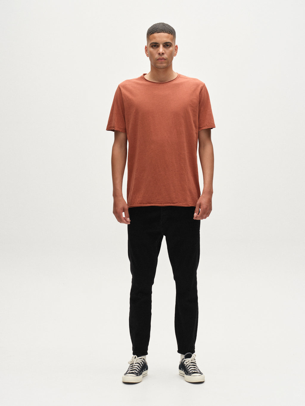 A person stands facing forward against a plain white background, wearing the Gabba Kostant New SS t-shirt in brown rust, paired with black pants and black-and-white sneakers.
