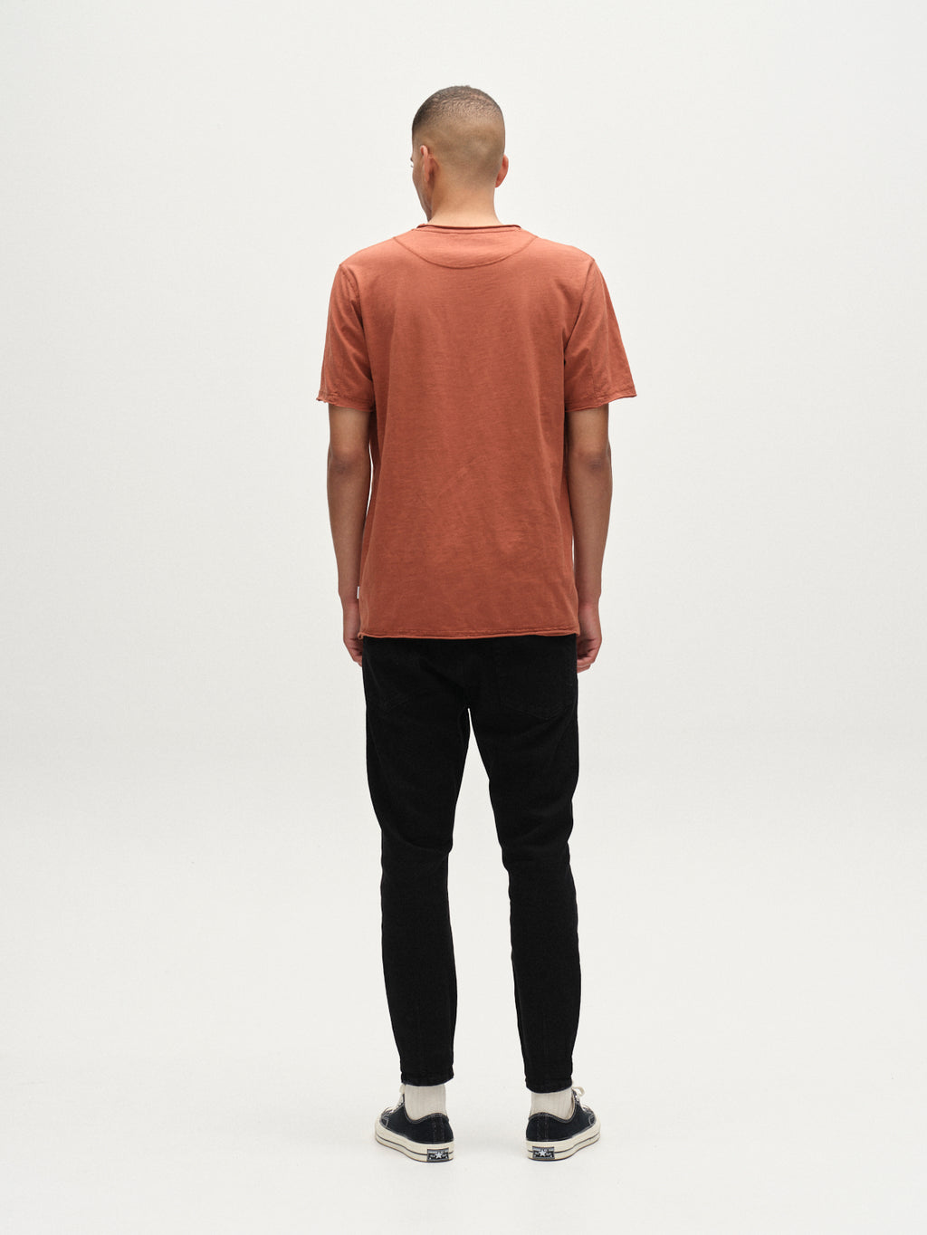 A person stands with their back to the camera wearing the Gabba Kostant New SS shirt in brown rust, paired with black pants and slip-on shoes, set against a plain white background.
