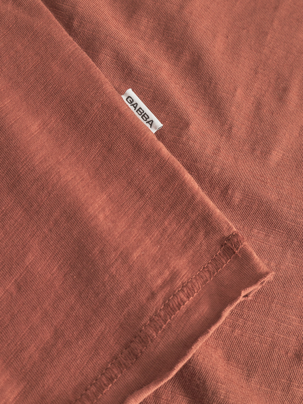 A close-up of the Gabba Kostant New SS T-shirt in brown rust, featuring breathable cotton, a small white Gabba tag, and visible stitching along the hem.